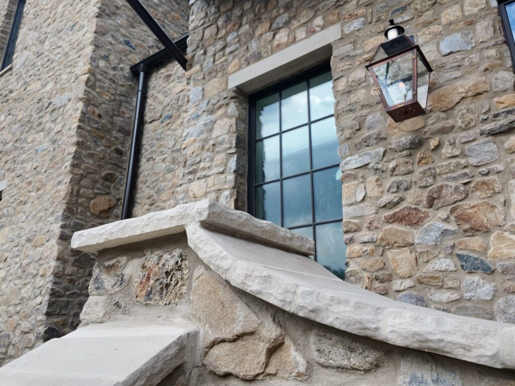 A detailed shot of a building's exterior masonry made of multi-toned natural stone. A prominent, smooth light-tan stone corbel with a curved profile is mounted on a corner. To the right, a black-framed multi-pane window reflects a cloudy blue sky. A traditional copper-and-glass lantern is mounted to the stone wall next to the window, featuring a warm-toned internal bulb. A black downspout is visible on the far left edge of the stone wall.