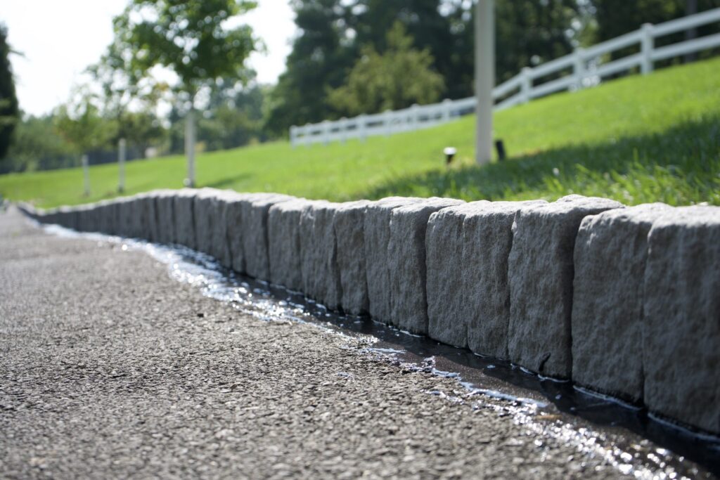 A close-up, low-angle shot of a gray asphalt driveway bordered by a row of light gray, rough-hewn stone pavers. The pavers have a jagged, natural texture.