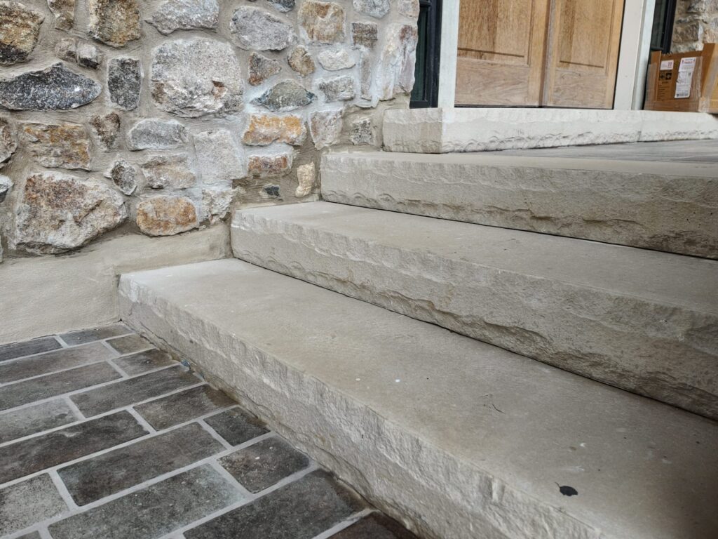 A ground-level, side-angle view of three heavy, light-tan natural stone steps. The steps feature a "chiseled" or "rock-faced" front edge and lead toward a set of wood doors. The staircase is built against a rustic stone veneer wall. The landing at the base of the stairs is paved with dark, rectangular cobblestone-style pavers.